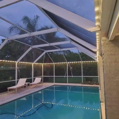 Dusk view: LED lights outlining screened pool with lounge chairs. Miami handyman installed LED string lights around screened pool enclosure.